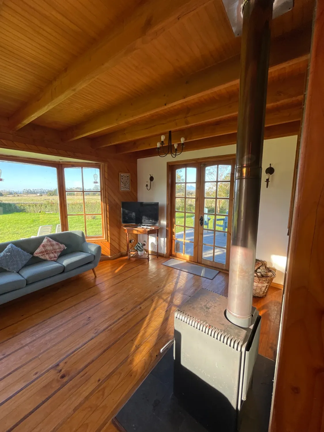 Living de la casa con estufa a leña, sofá y ventanales hacia el campo